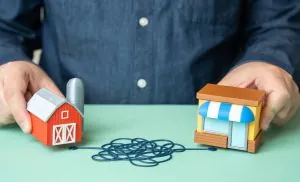 A visual metaphor of a person's hands holding a farm model and a retail storefront model separated by a chaotic, tangled line, representing the distribution challenges that strategic logistics leadership must solve.